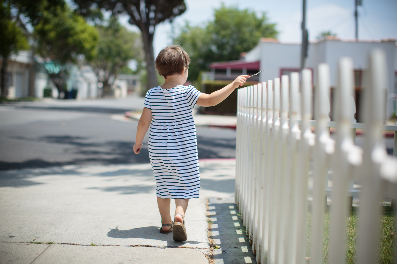 Little toddler girl in summer dress walking away along white fence. Back view.