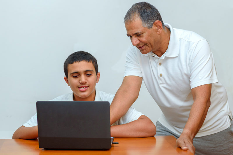 Senior Father and teenage son using laptop. Boy and dad sitting at home working with tablet computer. Happy family old grandfather and grandson on laptop. Elderly teacher trainer and teen pupil boy.