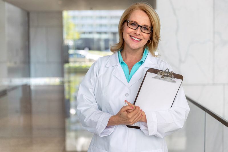 doctor physician, healthcare professional portrait, smiling sincere with clipboard at hospital clinic