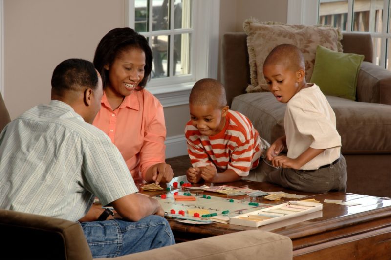 An African-American family (adult male and female and two male children) sit around a coffee table playing a board game