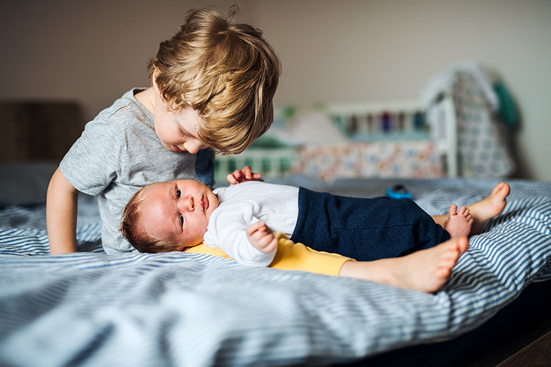 A small toddler boy with a newborn baby brother at home, sitting on bed.