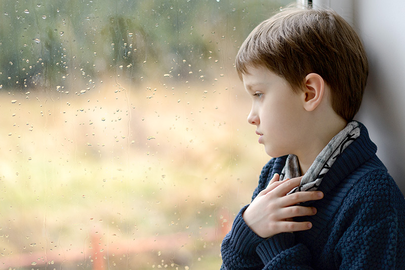 depressed and thoughtful little boy looking through the window on a rainy day