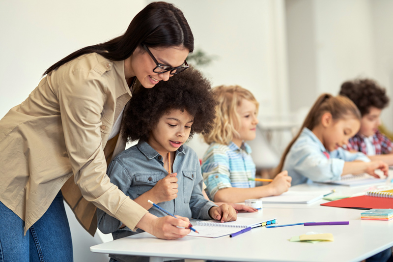 Female teacher helping a young student sitting at the table in elementary school classroom